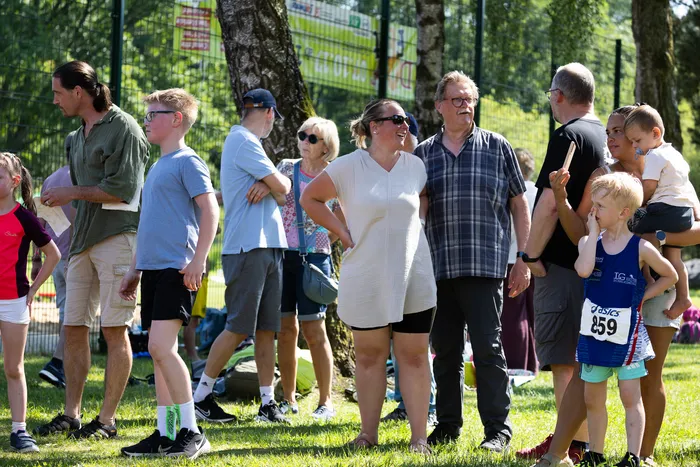 Eine Gruppe von Menschen steht an einem sonnigen Tag an der Seite einer Laufstrecke. Einige Zuschauer beobachten das Rennen, während ein junger Läufer mit der Startnummer 859 im Vordergrund steht.