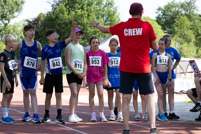 Ein Trainer in rotem Shirt erklärt einer Gruppe von Kindern und Jugendlichen in Sportkleidung etwas. Die Kinder tragen Startnummern und stehen auf einer Laufbahn.