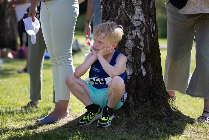 Ein kleiner Junge sitzt auf dem Gras am Stamm eines Baumes, den Kopf in die Hand gestützt. Er trägt ein blaues Tanktop, grüne Shorts und gelbe Turnschuhe. Im Hintergrund stehen Menschen auf einer Wiese.
