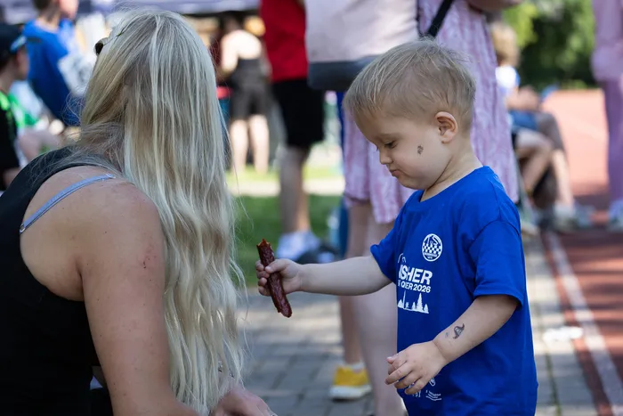 Ein kleiner Junge in blauem T-Shirt mit Aufschrift „Fischer Skier 2026“ reicht einer Person eine Wurst. Im Hintergrund sind weitere Menschen und Bäume zu sehen.