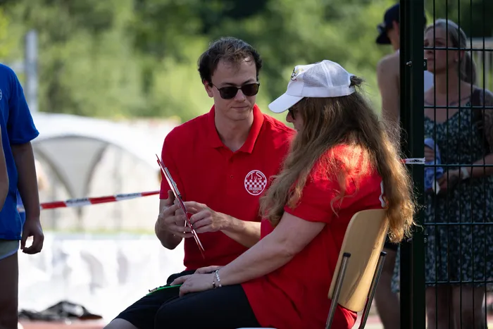 Ein Mann in rotem Poloshirt und eine Frau mit langen, blonden Haaren sitzen sich auf einer Sportanlage gegenüber und besprechen etwas auf einem Tablet. Im Hintergrund sind weitere Personen und ein Tennisfeld zu sehen.