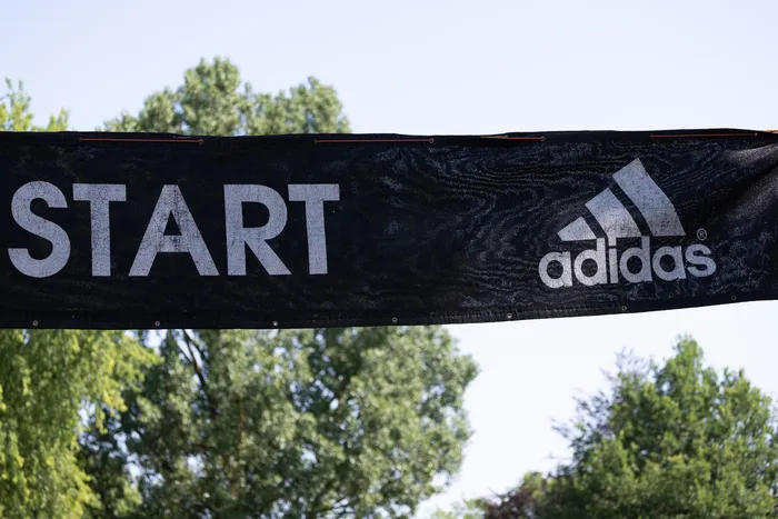 Schwarzes Startbanner mit weißer Schrift und dem Adidas-Logo, gespannt vor Bäumen und blauem Himmel.