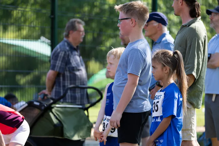 Junge und Mädchen in blauen Trikots mit Startnummern stehen nebeneinander und schauen in die Ferne. Im Hintergrund stehen weitere Personen und ein Kinderwagen.