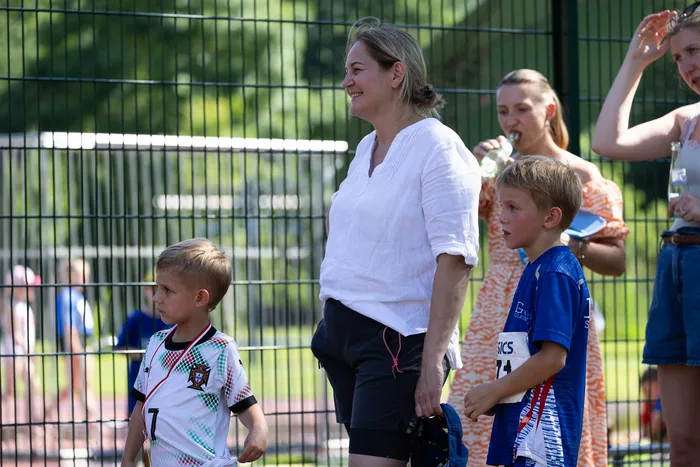 Eine Frau in weißer Bluse und dunkler Hose steht neben zwei Kindern in Fußballtrikots auf einer Sportanlage. Im Hintergrund sind weitere Personen und ein grüner Zaun zu sehen.