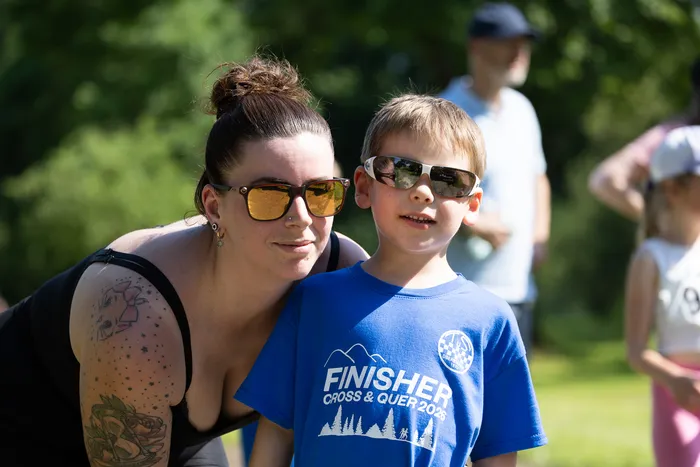 Eine Frau mit hochgestecktem Haar und Sonnenbrille steht neben einem Jungen, der ebenfalls eine Sonnenbrille trägt und ein blaues T-Shirt mit der Aufschrift 