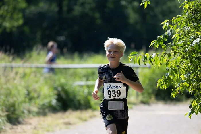 Lächelnder Junge mit blonden Haaren läuft bei einem Crosslauf, trägt eine Startnummer mit der Aufschrift 939 und Sportkleidung von Asics und Adidas. Im Hintergrund sind weitere Läufer und Bäume zu sehen.
