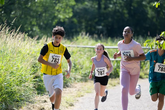 Vier Kinder laufen bei einem Crosslauf im Freien. Die Läufer tragen Startnummern und Sportkleidung, während sie auf einem Feldweg unterwegs sind.