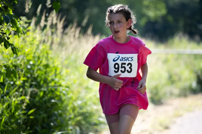 Ein junges Mädchen läuft bei einem Crosslauf, trägt ein pinkes T-Shirt mit der Startnummer 953 und Shorts. Sie blickt entschlossen nach vorne, während sie auf einem Waldweg unterwegs ist.