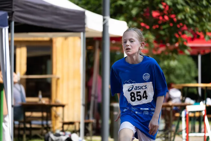 Ein junges Mädchen in einem blauen Trikot mit der Startnummer 854 rennt bei dem Cross- und Querlauf. Im Hintergrund sind Zuschauer und ein hölzerner Pavillon zu sehen.