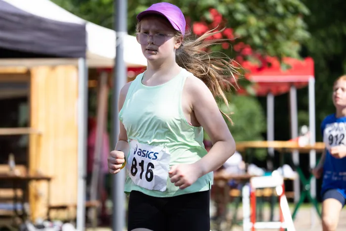 Eine Läuferin mit der Startnummer 816 in einem hellgrünen Top und schwarzer Laufhose rennt bei dem Cross- und Querlauf 2026. Ihr langes, blondes Haar weht im Wind. Im Hintergrund sind weitere Läufer und Zuschauer zu sehen.