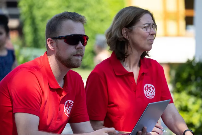 Zwei Personen mit roten Poloshirts sitzen nebeneinander, der Mann trägt eine Sonnenbrille und blickt zur Seite, die Frau schaut geradeaus. Beide halten ein Laptop oder Tablet in den Händen.