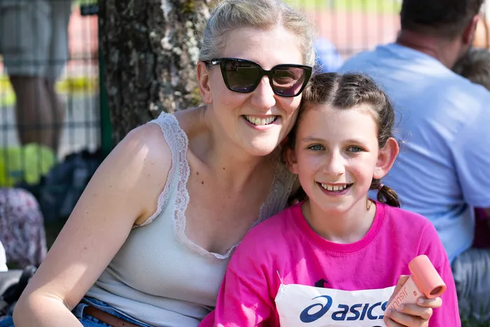 Eine Frau mit Sonnenbrille und ein lächelndes Mädchen stehen eng zusammen. Das Mädchen hält eine orangefarbene Trinkflasche, beide tragen Sportkleidung.