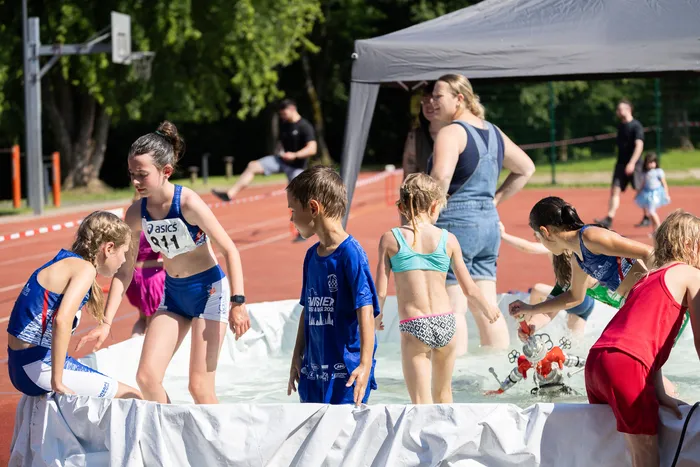 Mehrere Kinder und Jugendliche stehen in einem flachen Wasserbecken auf einer roten Tartanbahn. Im Hintergrund steht eine erwachsene Person und weitere Sportler sind zu sehen.