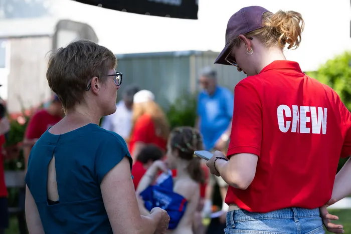 Zwei Personen stehen einander zugewandt, eine trägt ein rotes T-Shirt mit der Aufschrift „CHEW“, die andere ein blaues Oberteil. Im Hintergrund sind weitere Menschen und Zelte auf einem Festivalgelände zu sehen.