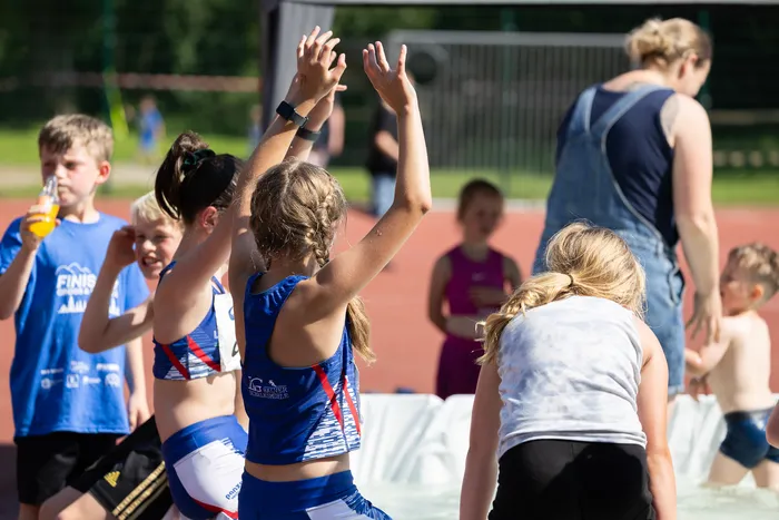 Junge Athleten mit erhobenen Händen stehen auf einer roten Tartanbahn, während ein Wettbewerb stattfindet. Im Hintergrund sind weitere Kinder und Jugendliche sowie Zuschauer zu sehen.