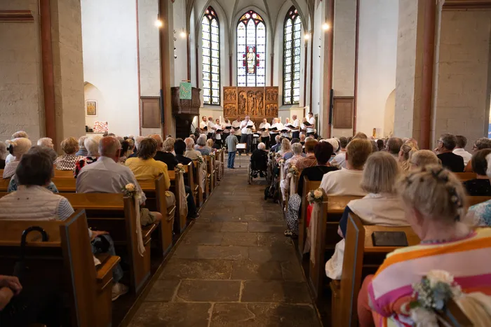 Ein Chor singt in einer Kirche vor einer versammelten Gemeinde. Die Kirchenbänke sind gefüllt mit Zuschauern, und durch die hohen Kirchenfenster fällt Licht in den Raum.