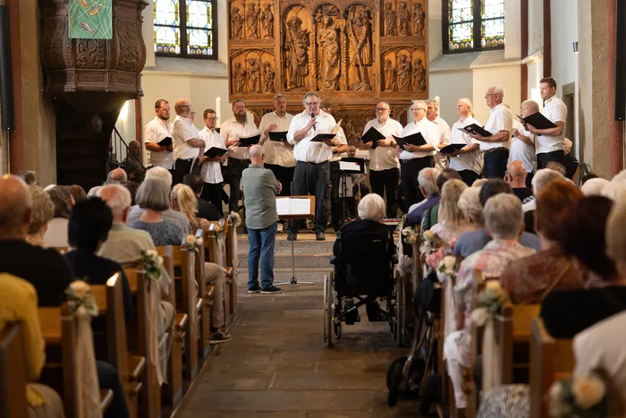 Ein Chor singt in einer Kirche vor einem Publikum. Der Dirigent steht vor dem Chor und hält die Partitur in der Hand.