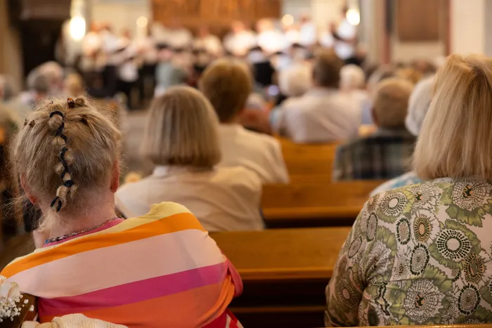 Zuschauer sitzen auf Holzbänken in einer Kirche und blicken auf eine Gruppe von Chorsängern, die im Hintergrund zu sehen sind. Im Vordergrund ist der Nacken einer Frau mit geflochtenem Haar und einem bunt gestreiften Oberteil zu erkennen.
