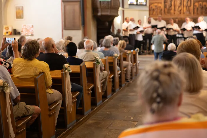 Chor singt in einer Kirche vor einem Publikum. Die Bänke sind gefüllt mit Zuschauern, einige halten ihre Smartphones hoch.