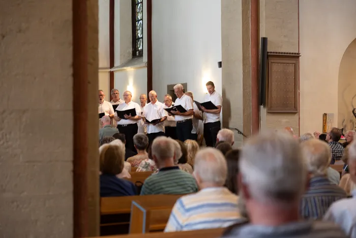 Ein Chor singt in einer hellen Kirche vor einem Publikum. Die Sänger, hauptsächlich ältere Männer, halten Notenblätter in der Hand.