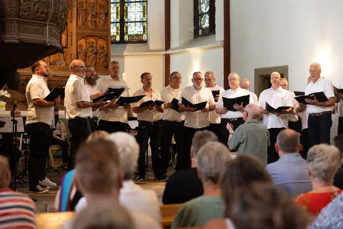 Ein Männerchor singt in einer Kirche, die Zuhörer sitzen auf Bänken im Vordergrund. Im Hintergrund ist ein Altar mit einer bunten Glasfenster zu sehen.