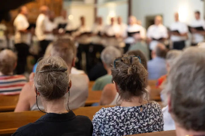 Zuschauer sitzen auf Holzbänken in einer Kirche und blicken auf eine Gruppe von Chorsängern, die vorne aufgestellt sind. Im Vordergrund sind die Hinterköpfe von Personen zu sehen.