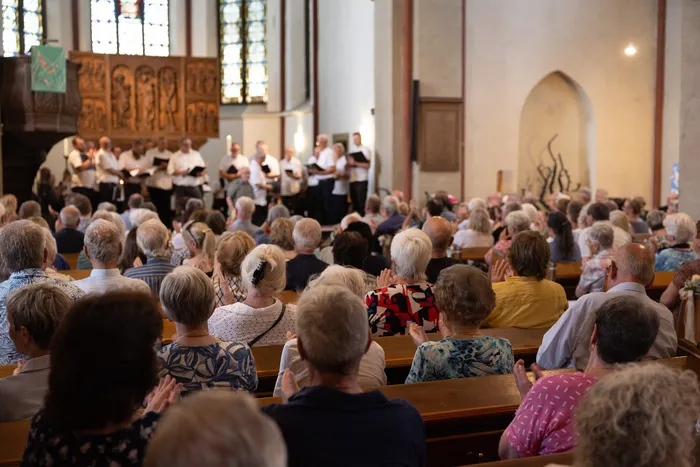 Ein Chor singt in einer Kirche vor einem Publikum, das hauptsächlich aus älteren Menschen besteht. Einige Zuhörerinnen und Zuhörer klatschen.