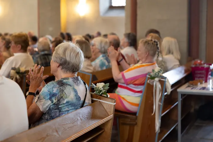 Zuschauer sitzen auf Kirchenbänken und klatschen während eines Konzerts in der Jakobus-Kirche. Im Vordergrund eine Frau mit kurzem, grauem Haar und ein blau gemustertes Oberteil.