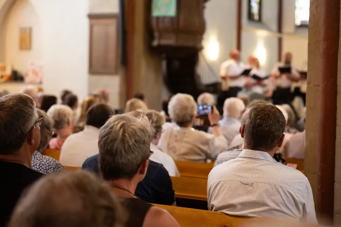 Ein Publikum sitzt auf Holzbänken in einer Kirche und schaut in Richtung einer Gruppe von Chorsängern, die Notenblätter halten. Im Hintergrund ist ein hölzerner Kirchenaltar zu sehen.