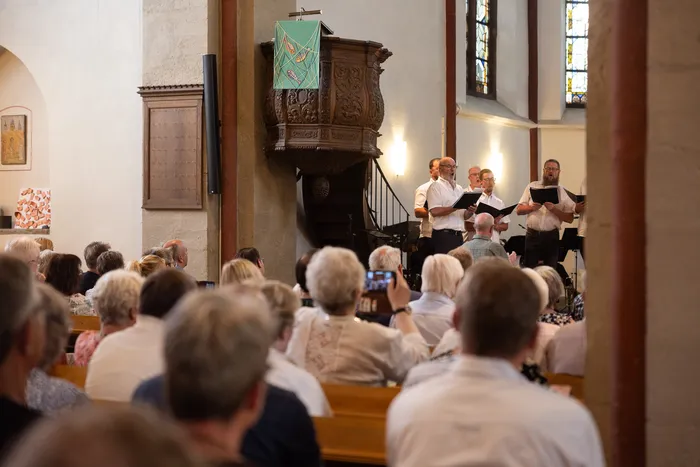 Ein Männerchor singt vor einer versammelten Gemeinde in der Jakobus-Kirche. Die Zuhörer sitzen auf Holzbänken und verfolgen den Auftritt.