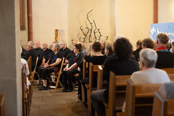 Eine Gruppe von Menschen sitzt auf Holzbänken in einer Kirche und blickt nach vorne. Im Hintergrund befindet sich eine abstrakte Metallskulptur an der Wand.