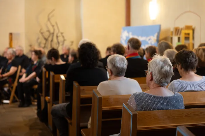 Menschen sitzen auf Kirchenbänken in einer Kirche und schauen in Richtung eines nicht sichtbaren Vordergrunds, vermutlich zu einer Bühne. Im Hintergrund ein Banner und eine abstrakte Skulptur.