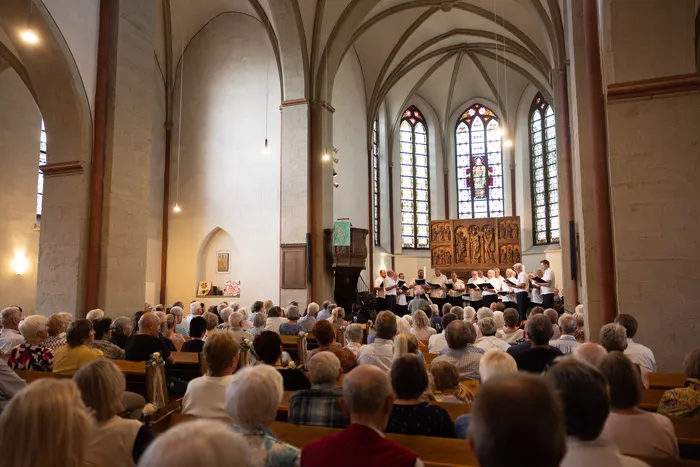 Ein Chor singt in einer hellen Kirche vor einem Publikum, das auf Bänken sitzt. Bunte Glasfenster erhellen den Raum.