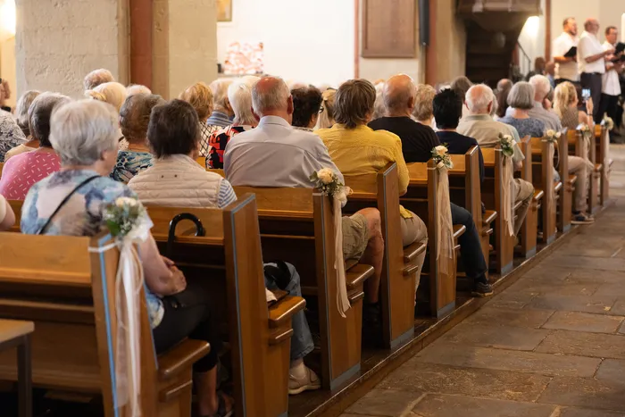 Zuschauer sitzen auf Holzbänken in einer Kirche und hören einem Chor zu. Die Bänke sind mit Schleifen und Blumen geschmückt.