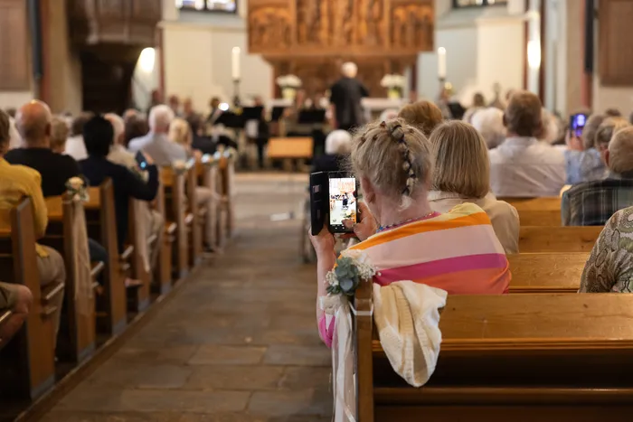 Zuschauer filmen mit einem Smartphone während eines Konzerts in einer Kirche. Die hölzernen Kirchenbänke sind fast vollständig gefüllt.