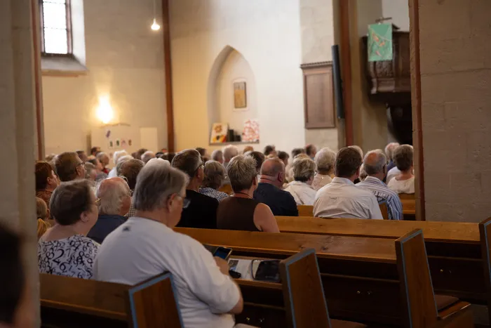 Eine Menschenmenge sitzt auf Holzbänken in einer Kirche und blickt nach vorne. Im Hintergrund ist ein Altar und weitere Kirchenelemente zu sehen.
