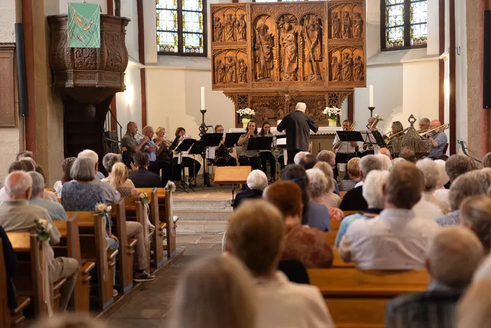 Ein Orchester spielt vor einem Publikum in einer Kirche. Im Hintergrund ist ein reich verzierter Altar mit religiösen Figuren zu sehen.