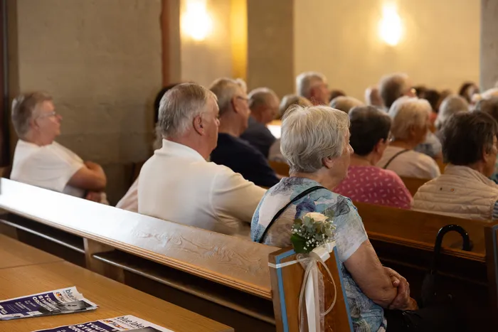 Zuschauer sitzen auf Holzbänken in einer Kirche und verfolgen ein Konzert. Im Vordergrund eine Frau mit grauen Haaren und eine Brosche.