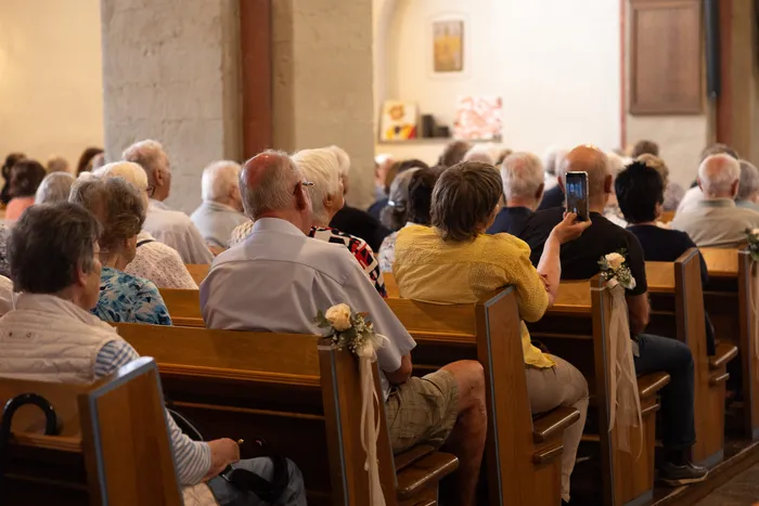 Zuschauer sitzen auf Holzbänken in einer Kirche und hören einem Konzert zu. Eine Frau filmt mit ihrem Smartphone.