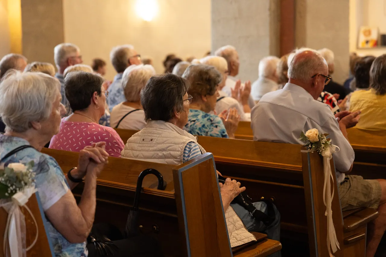Eine Gruppe älterer Menschen sitzt auf Holzbänken in einer Kirche und lauscht einem Konzert. Einige haben die Hände gefaltet oder klatschen.