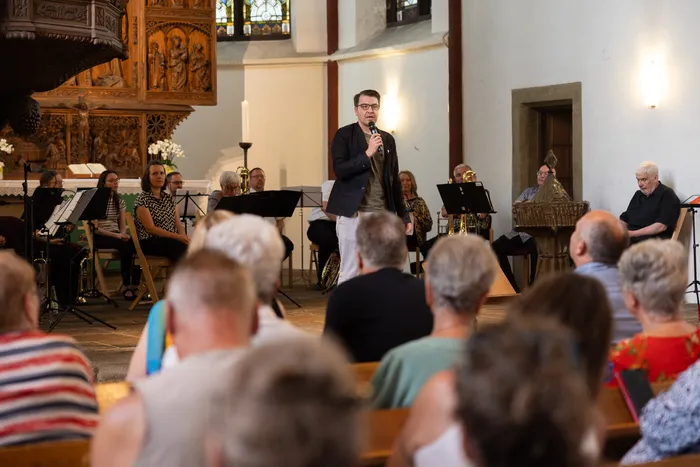 Ein Mann mit Brille spricht in ein Mikrofon vor einem Ensemble von Musikern und einem Publikum in einer Kirche. Im Hintergrund ist ein kunstvoll geschnitzter Altar zu sehen.