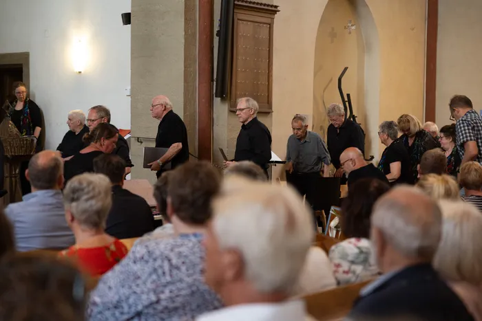 Ein Chor singt in einer Kirche vor einem Publikum. Mehrere Sänger stehen mit Notenblättern vor Mikrofonen, während das Publikum auf Bänken sitzt.