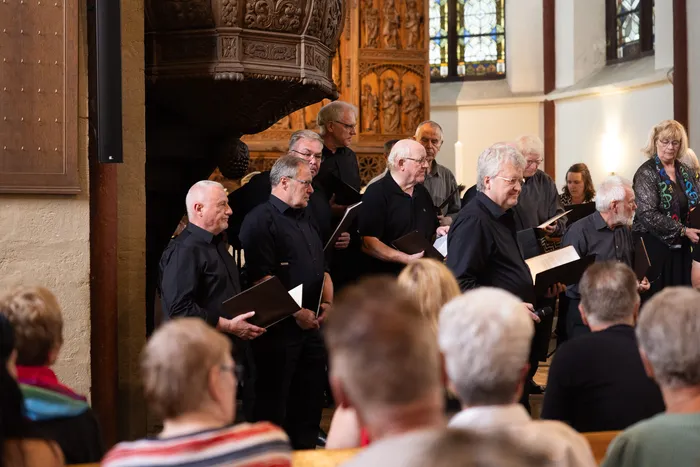 Ein Chor singt in einer Kirche, die Mitglieder halten Notenblätter in der Hand. Im Hintergrund sind weitere Personen und Kirchenfenster zu sehen.