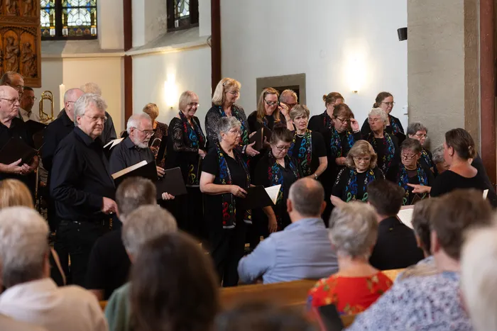 Ein Chor singt in einer Kirche, die Zuhörer sitzen auf Bänken im Vordergrund. Die Chormitglieder halten Notenblätter in der Hand und tragen dunkle Oberteile mit farbenfrohen Mustern.