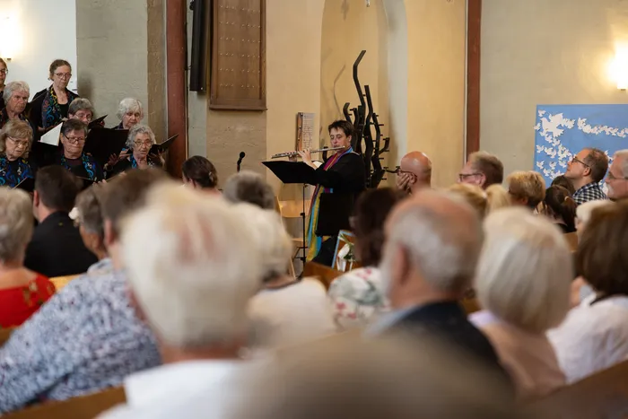 Eine Frau mit kurzen Haaren und schwarzem Oberteil dirigiert einen Chor in einer Kirche. Im Vordergrund sitzen oder stehen zahlreiche Zuschauer und verfolgen die Aufführung.
