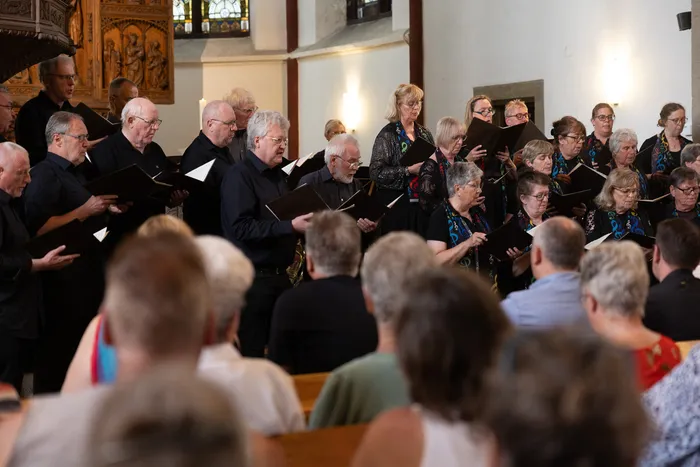 Ein Chor singt in einer Kirche, während im Vordergrund Zuschauer sitzen. Die Sängerinnen und Sänger halten Notenblätter in den Händen.