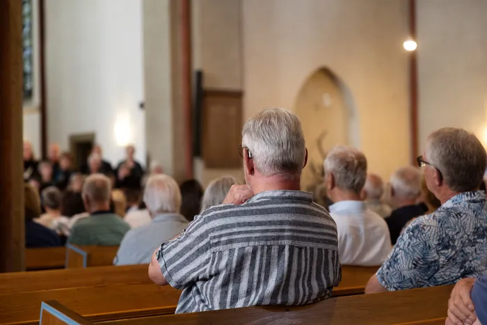 Menschen sitzen auf Holzbänken in einer Kirche und schauen in Richtung einer Gruppe, die weiter vorne steht. Im Hintergrund ist ein Bogen zu sehen.