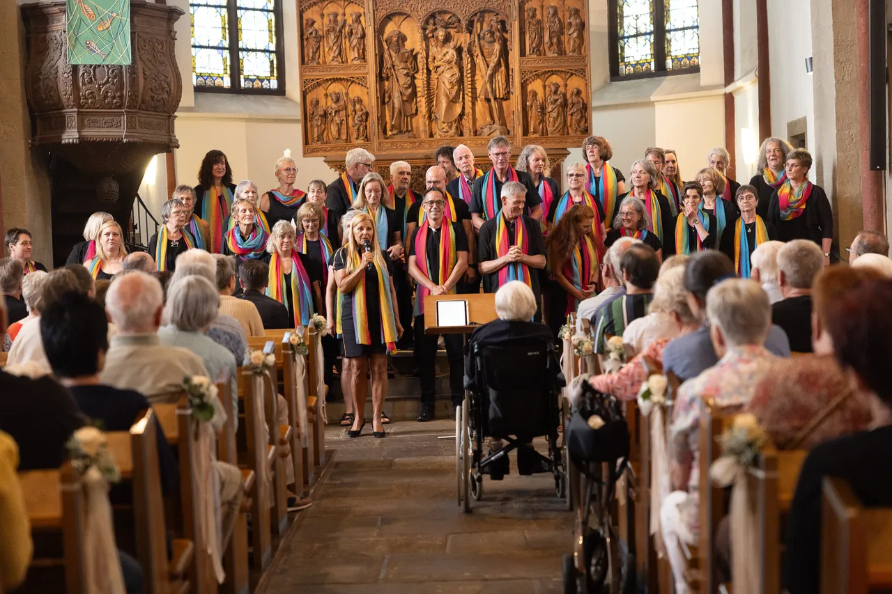 Ein Chor steht auf der Empore einer Kirche und singt vor einem Publikum. Die Chormitglieder tragen bunte Tücher und stehen vor einem Altar mit religiösen Figuren. Einige Zuschauer sitzen in Kirchenbänken, eine Frau sitzt im Rollstuhl.