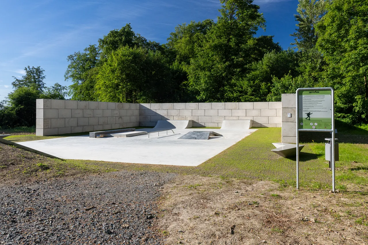 Betonierter Skatepark mit verschiedenen Rampen und Hindernissen, umgeben von Grünflächen und Bäumen. Ein Schild mit Regeln und Informationen steht am Rand.