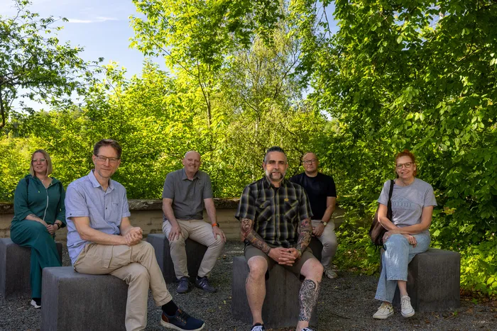Sechs Personen sitzen im Freien auf grauen Betonblöcken vor üppigem Grün. Sie spenden 2500 Euro für Obdachlosenhilfe.